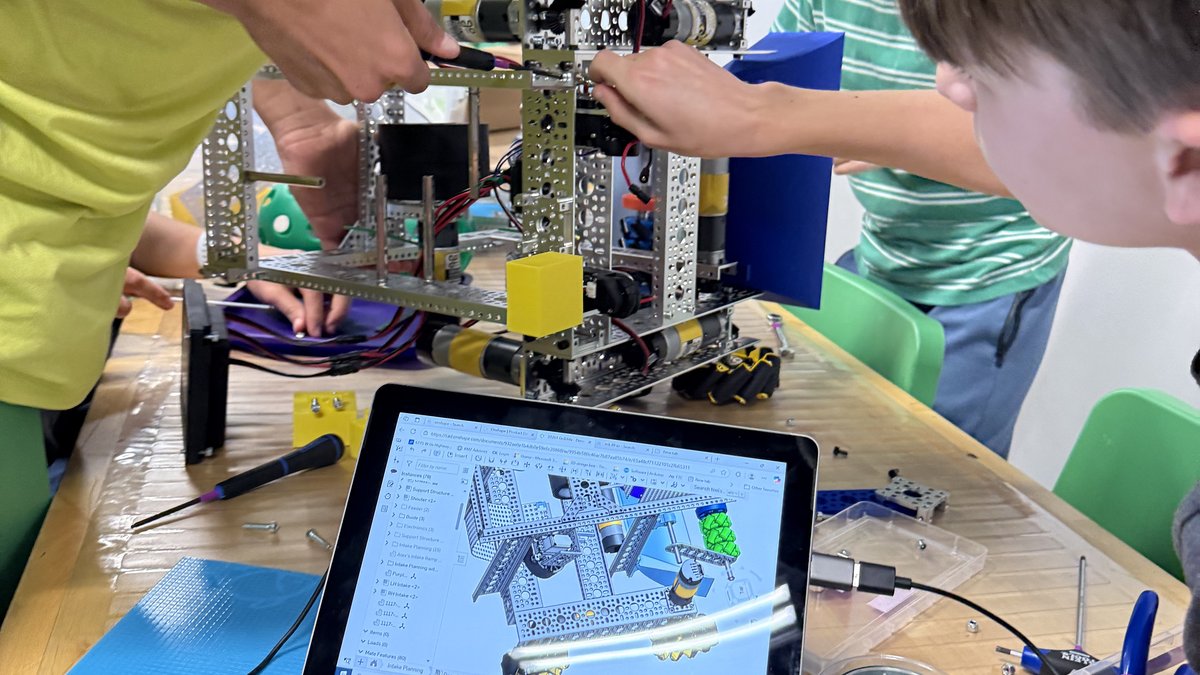 Students assemble robot components at a workbench while a CAD model displays on a laptop.