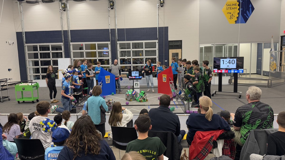 Wide view of a FIRST Tech Challenge competition floor, with two alliances of drive teams and spectators watching.